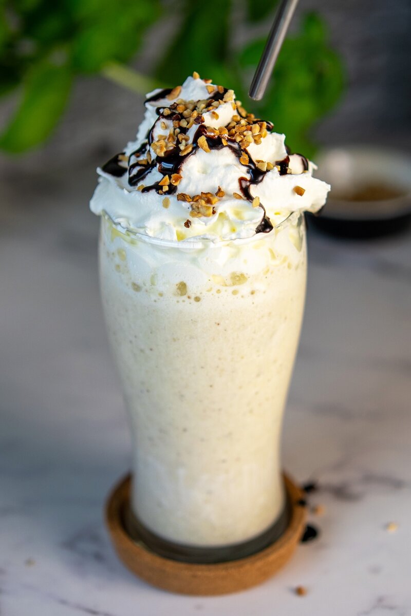 Creamy banana milkshake with whipped cream, chocolate syrup drizzle, and crushed nuts, refreshing homemade smoothie in tall glass.