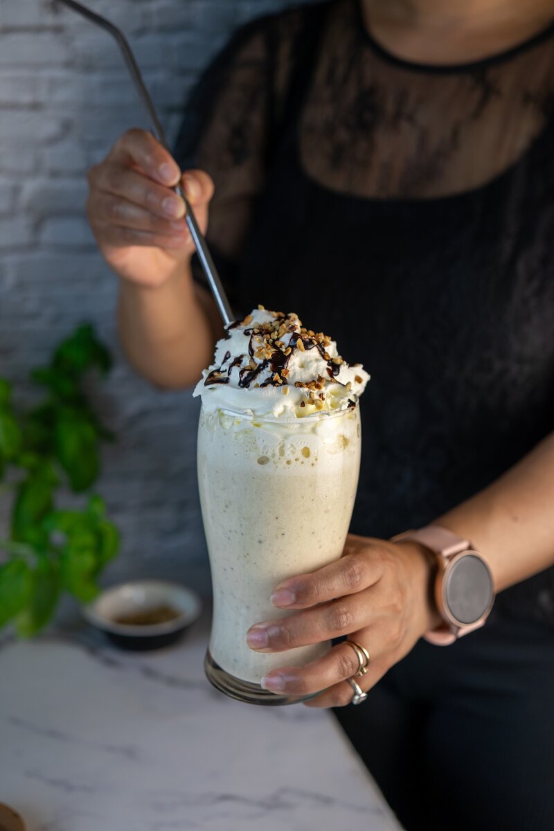 Person holding a tall banana milkshake with whipped cream, nuts, and chocolate syrup, handheld summer drink close‑up.