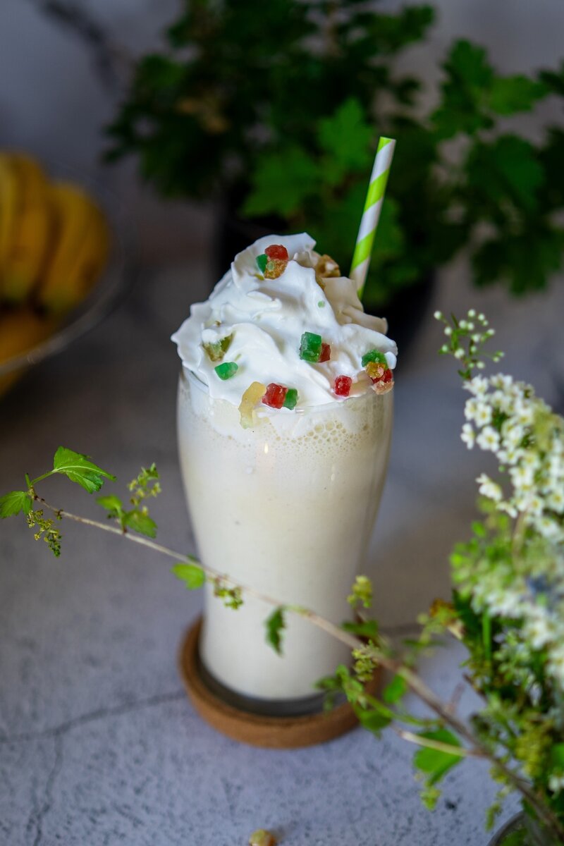 Tall banana milkshake in a chilled glass topped with whipped cream and colorful candied fruit, green striped straw, summer dessert.