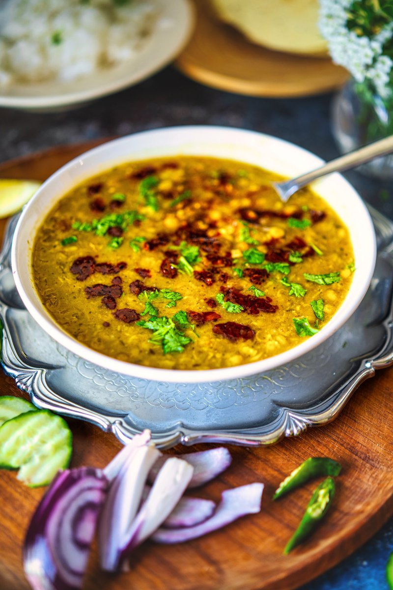 A bowl of chana dal garnished with cilantro and red chili tempering, served with cucumber slices, onion pieces, and green chilies on a wooden tray.