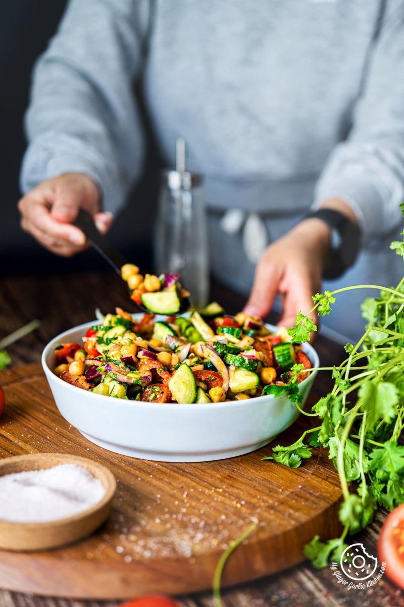 a female holding a black spoon filled with chickpea salad with one hand and chickpea salad bowl with other hand