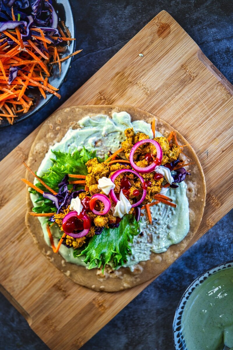Overhead view assembling chickpea tikka wrap with green yogurt sauce, crisp veggies, and pickled onions on whole wheat tortilla, vegan lunch idea