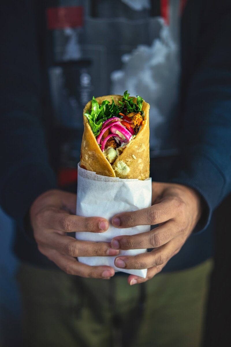 Hands holding a wrapped chickpea tikka vegan roll in paper, filled with lettuce, carrots, onions, and herb sauce, Indian-inspired healthy snack 