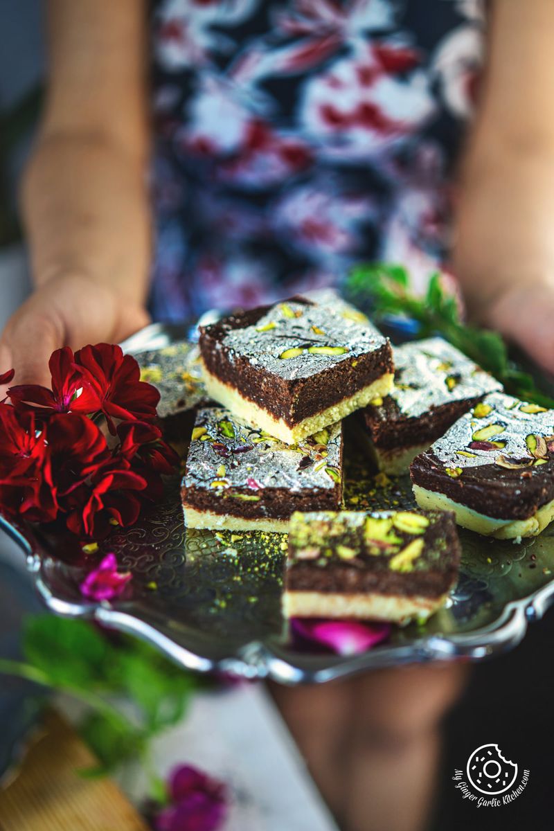 closeup shot of a female holding five chocolate burfis in a metal plate