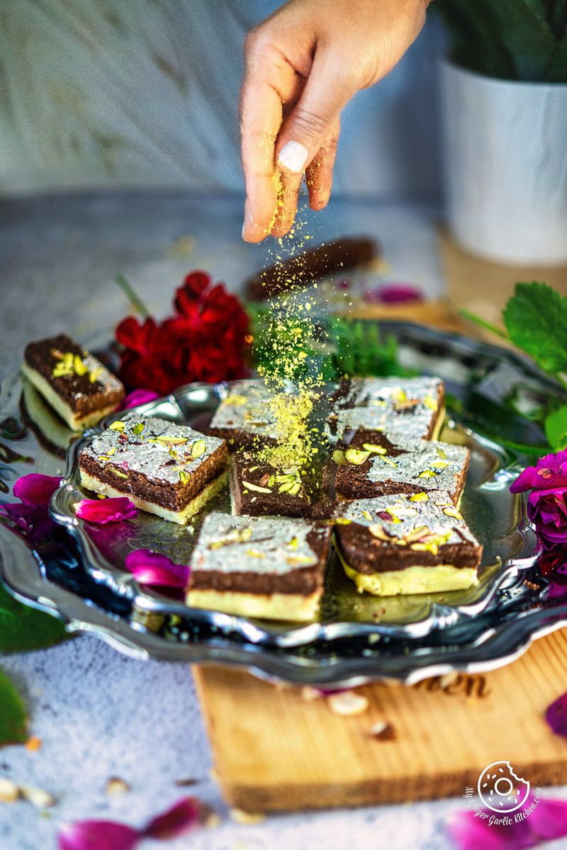 a hand sprinkling chopped pistachios over six chocolate burfis in a metal plate