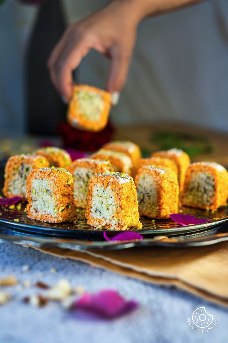 a female holding a burfi over coconut burfi rolls in a metal plate
