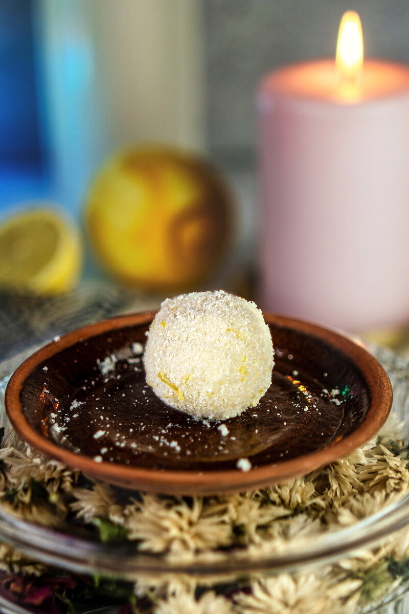 Close-up of a single white cottage cheese lemon ball dusted with coconut on a brown ceramic plate, with a pink candle and fresh lemons in the background