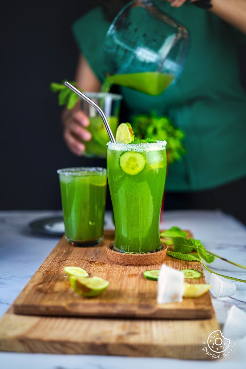 two glasses filled with cucumber mint cooler with a female pouring cooler in a glass