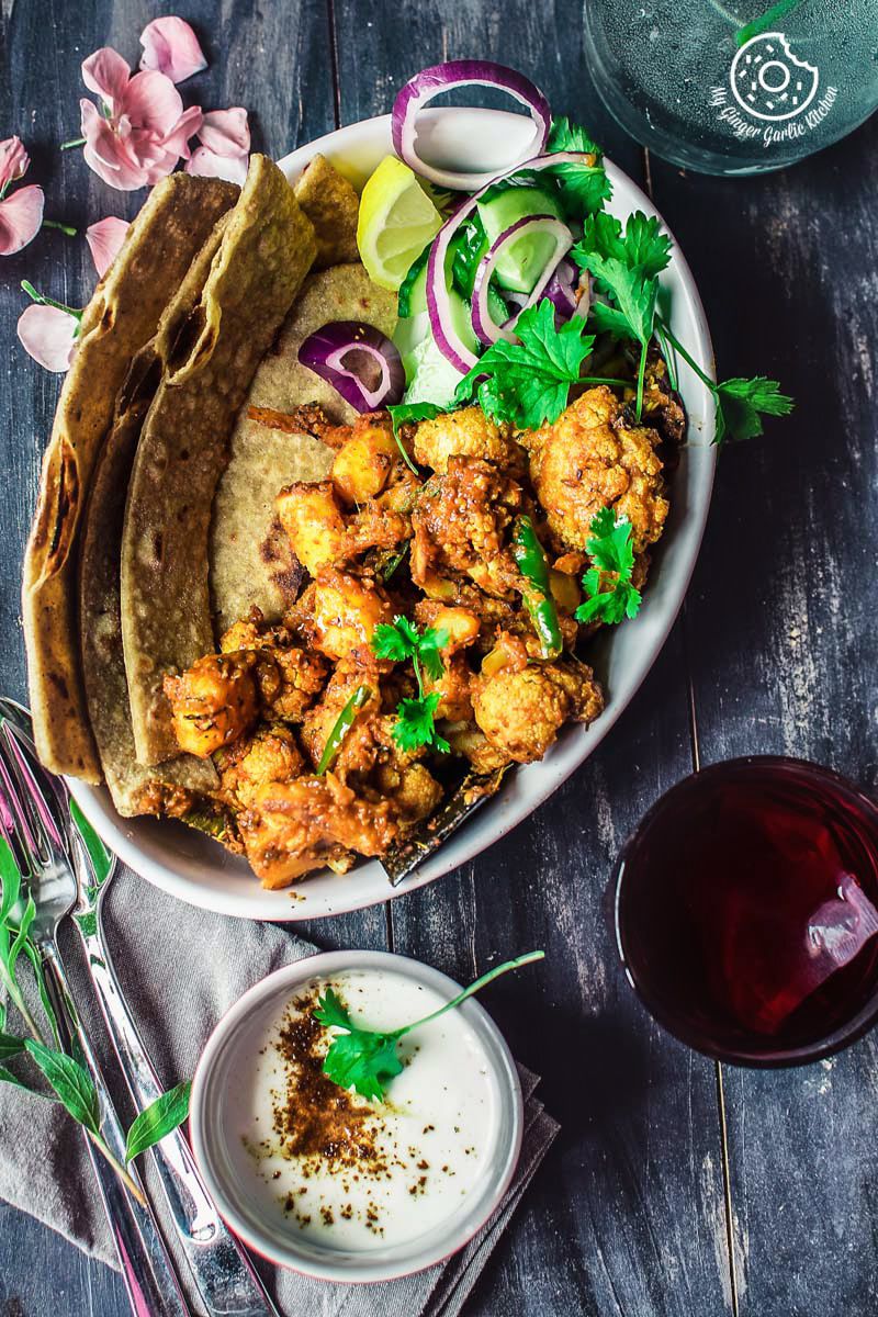 a plate of dhaba style dry aloo gobi with a paratha, onions, a bowl of yogurt, and a glass of tea