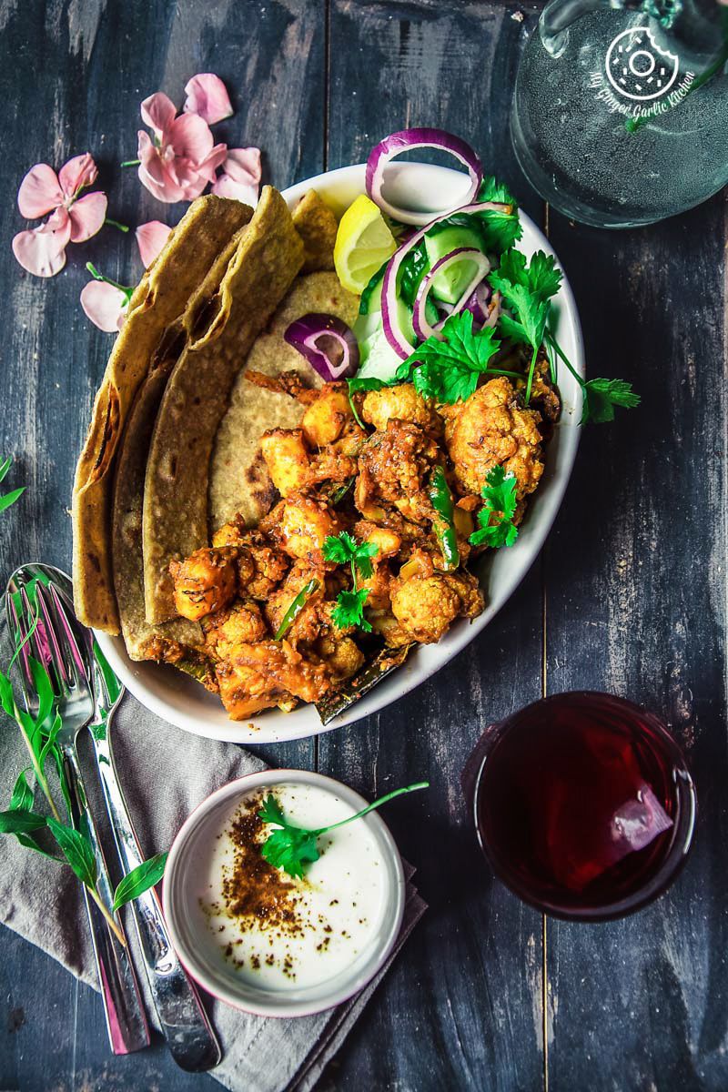 a close up of a plate of a plate of dhaba style dry aloo gobi with a paratha, onions, a bowl of yogurt, with a a glass of tea, flowers and fork on the side