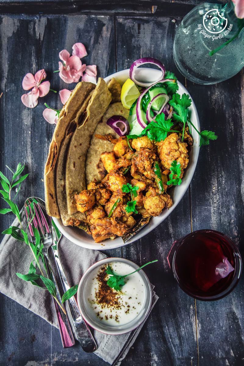 a close up of a plate of a plate of dhaba style dry aloo gobi with a paratha, onions, a bowl of yogurt, and a glass of tea 