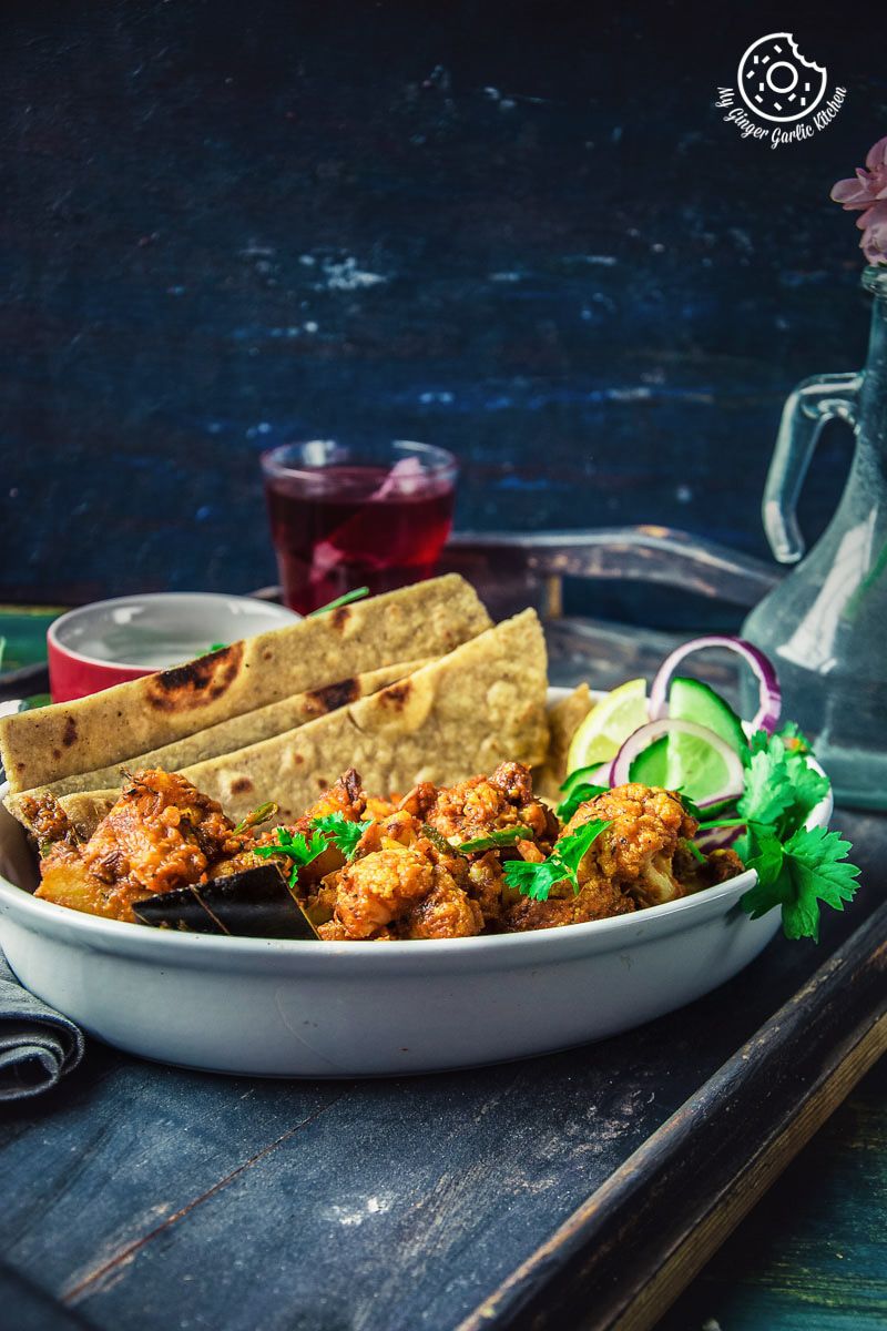 there is a bowl of a dhaba style dry aloo gobi with a paratha, onions, a bowl of yogurt, and a glass of tea