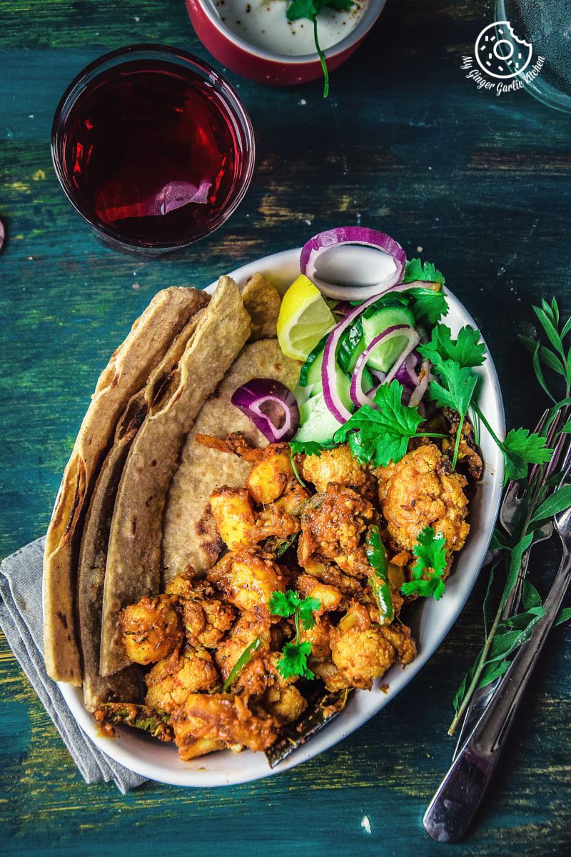 there is a plate of dhaba style dry aloo gobi with a paratha, onions, a bowl of yogurt, and a glass of tea