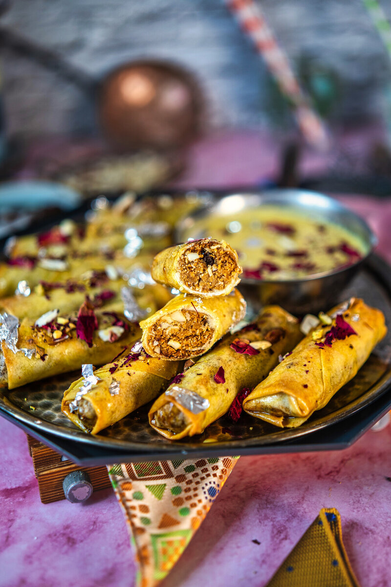 Cross-section view of crispy gujiya spring rolls showing the sweet filling inside, arranged on a black plate with rose petals and pistachio garnish