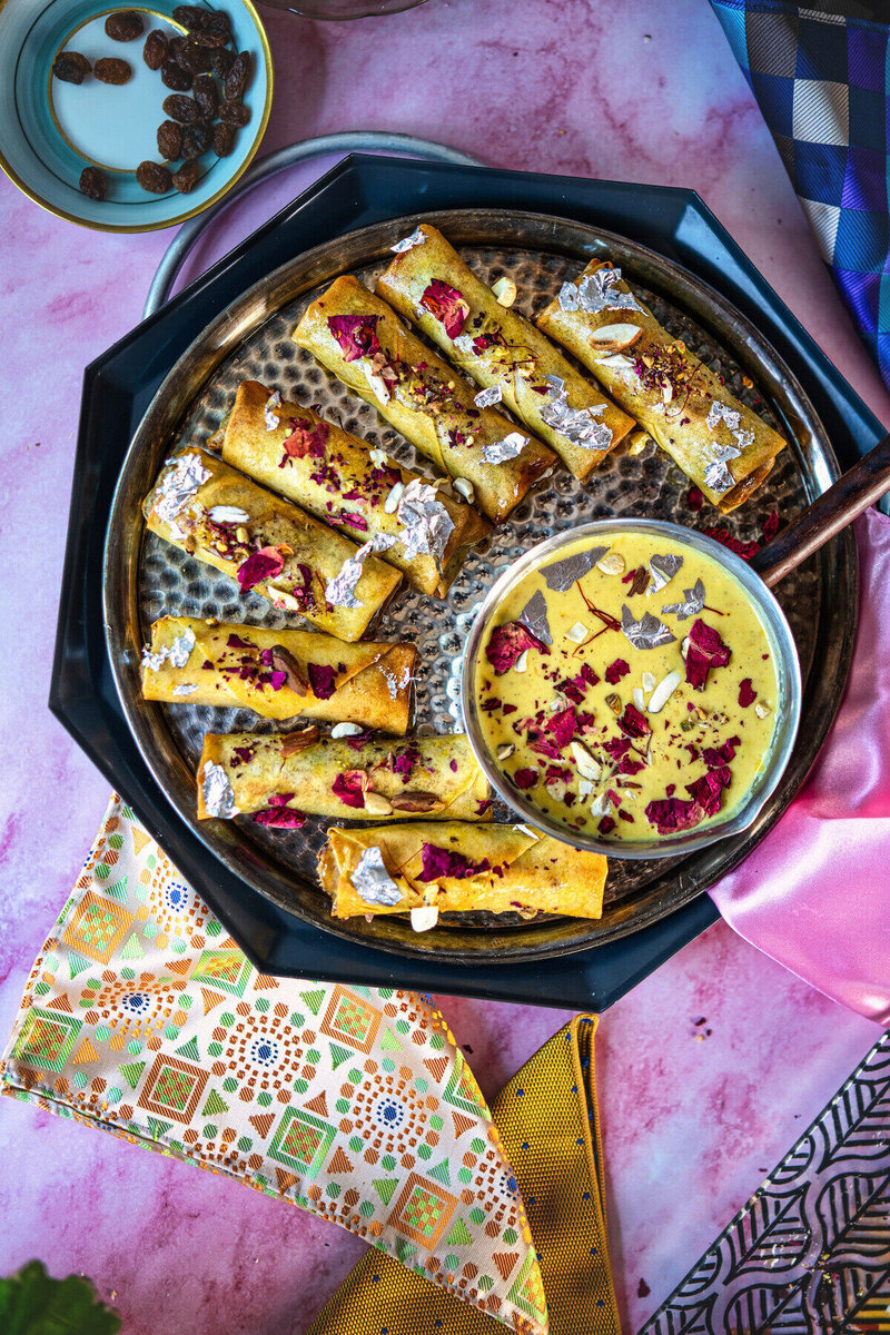 Crispy golden gujiya spring rolls arranged on a black platter, garnished with rose petals and pistachios, served with yellow dipping sauce on a pink background with decorative napkins