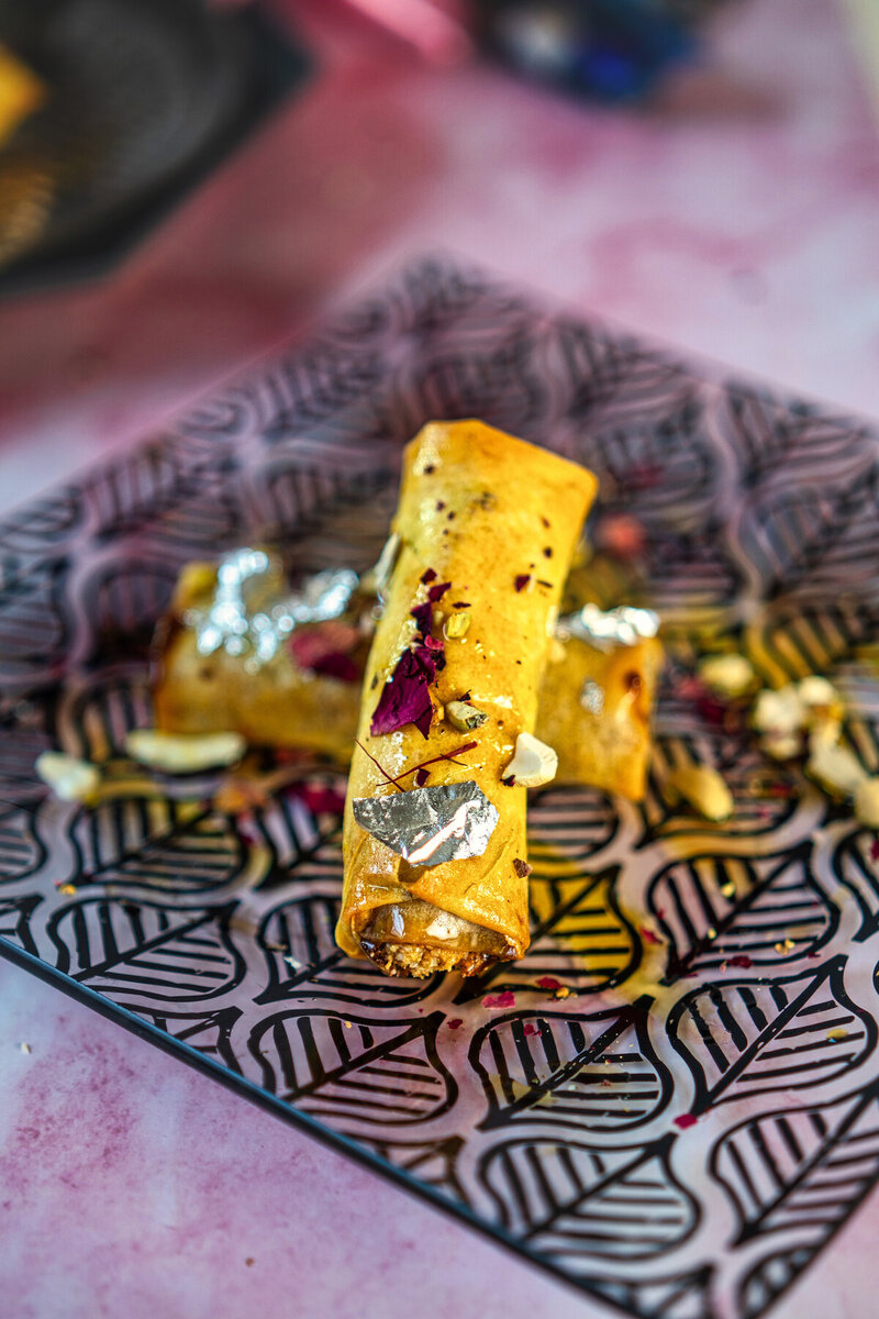 Close-up of a single gujiya spring roll on a patterned plate, showing crispy texture, silver foil decoration, and garnished with rose petals and nuts