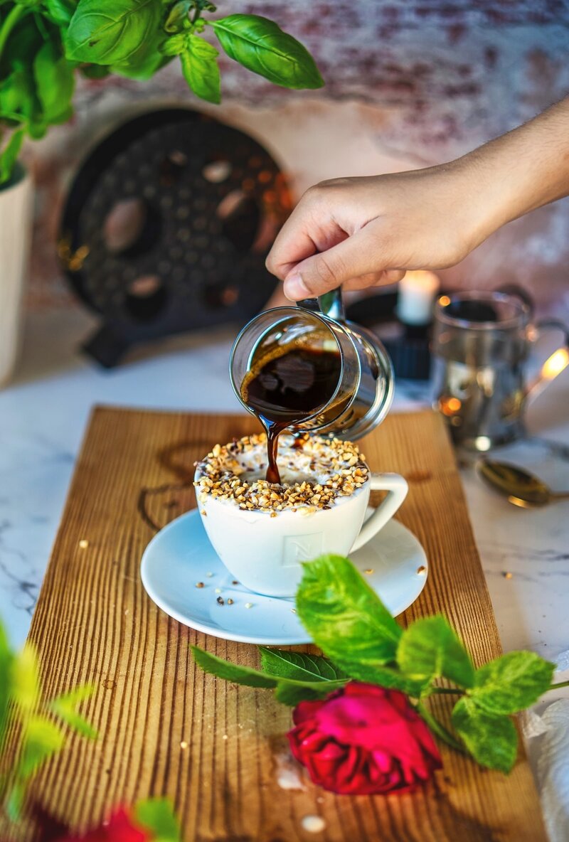 Hand pouring espresso over hazelnut affogato in white cup topped with chopped hazelnuts, surrounded by fresh red rose and green leaves