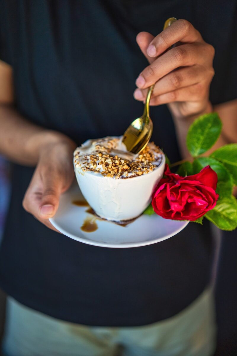 Person holding hazelnut affogato dessert in white cup with gold spoon topped with chopped hazelnuts and a red rose garnish