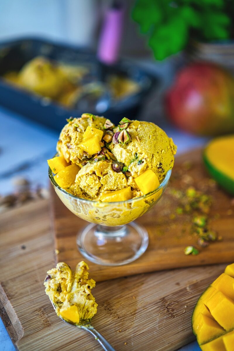 High protein mango ice cream being scooped from a loaf pan with an ice cream scooper, surrounded by diced mango pieces.