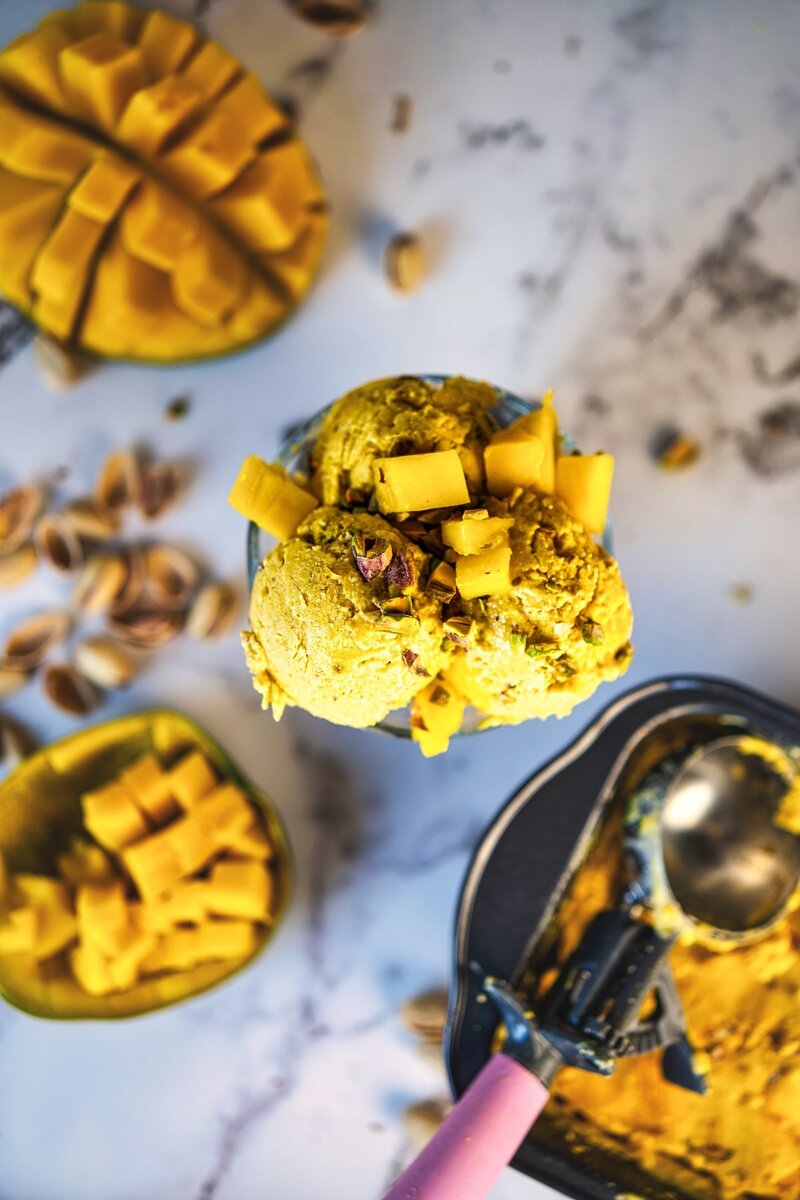 Overhead view of scoops of high protein mango ice cream in a glass, topped with chopped mango and nuts, with fresh mango and pistachios scattered on a marble surface.