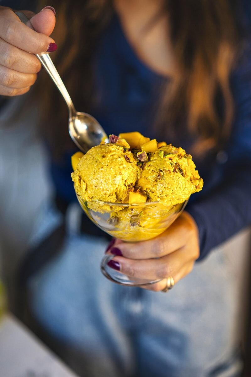 Person holding a glass bowl filled with scoops of high protein mango ice cream, garnished with diced mango and nuts, about to eat with a spoon.