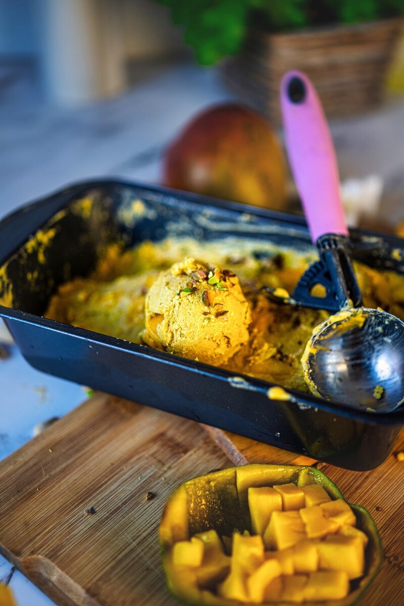 Glass bowl of high protein mango ice cream topped with diced mango and nuts, set on a wooden board with a spoonful of ice cream beside it, and fresh mangoes in the background.