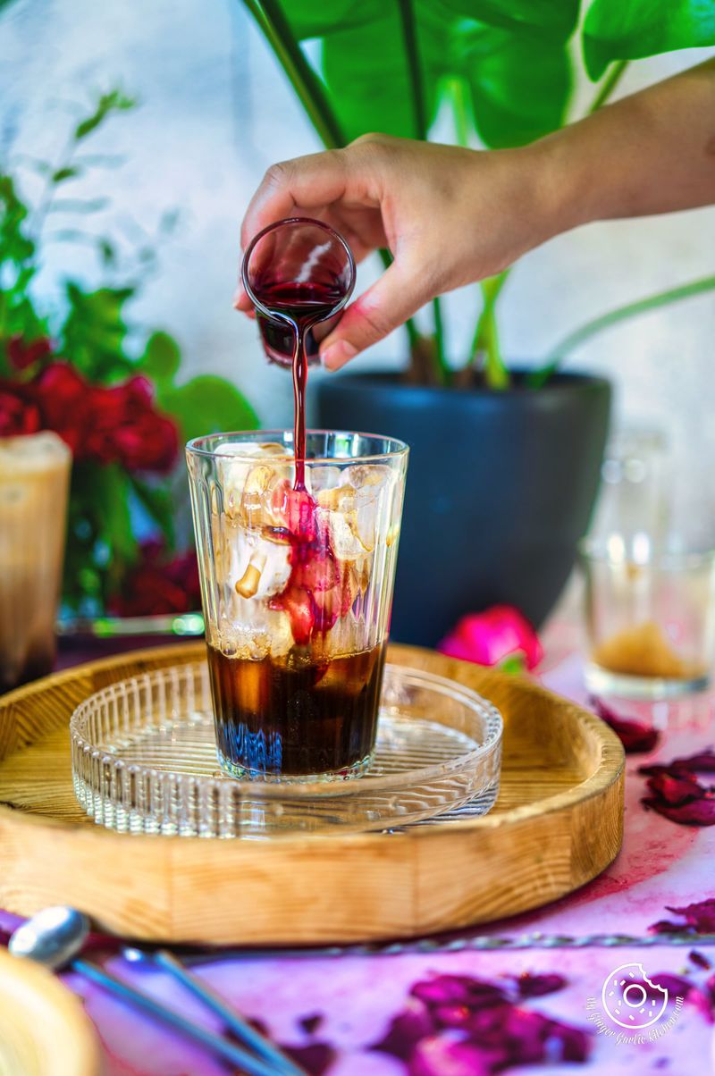 Picture of the iced rose latte. A hand is pouring rose syrup over ice cubes into the glass.
