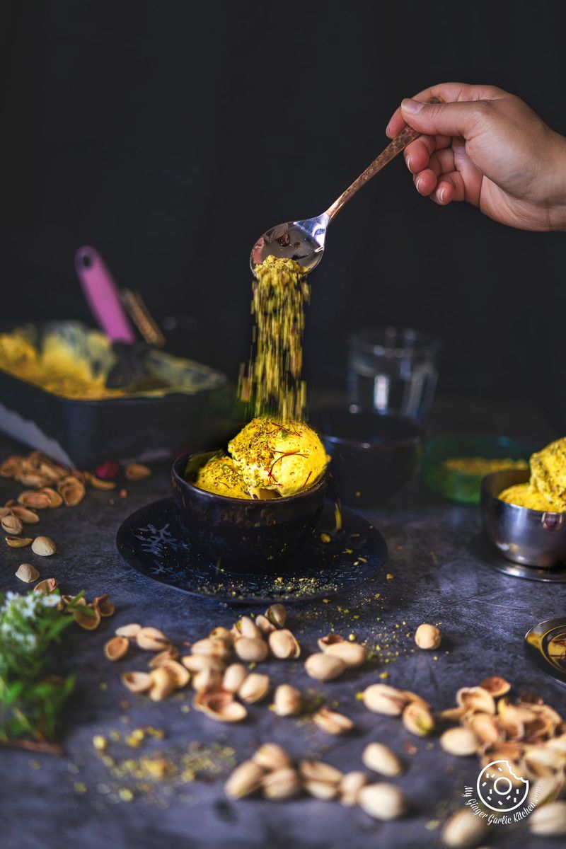 a hand sprinkling pistachio with a spoon over two kesar pista ice cream scoops in a small bowl