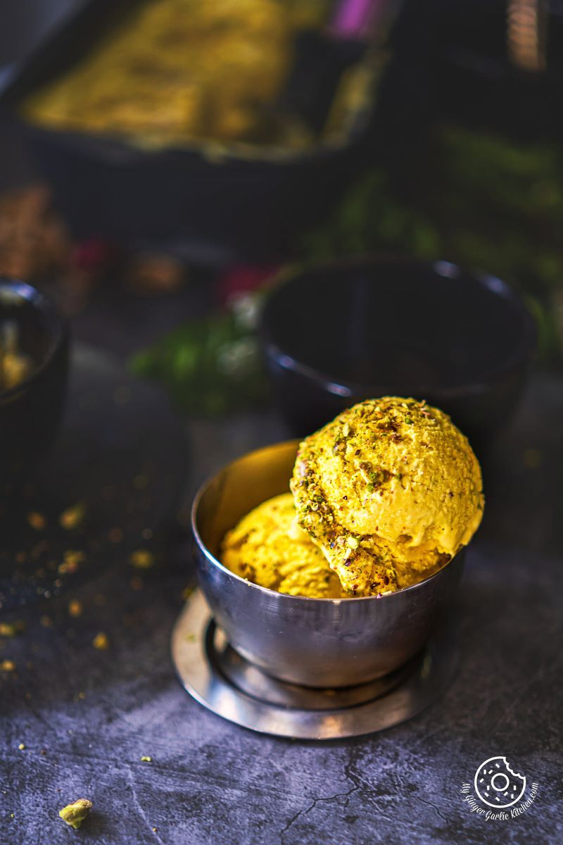 two kesar pista ice cream scoops in a metal bowl with a ice cream container cloth in the background
