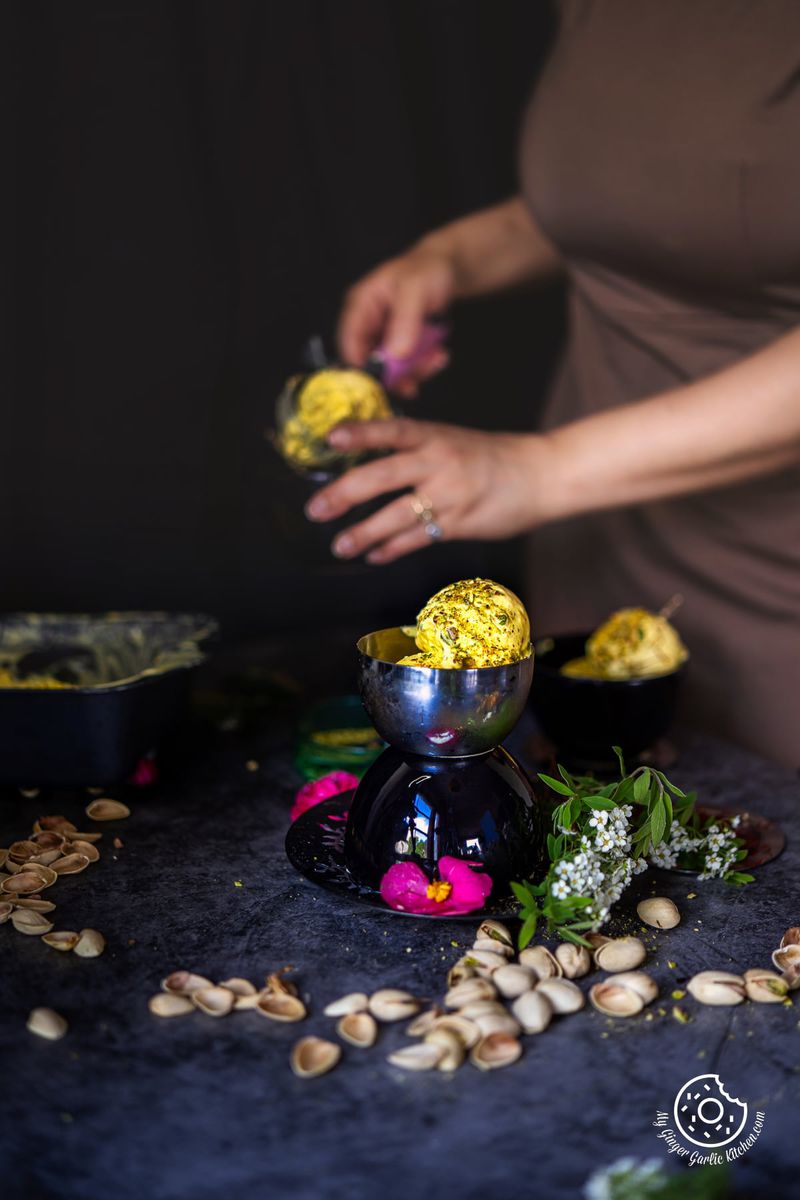 two kesar pista ice cream scoops in a small black bowl with a female holing an ice cream scoop in the background