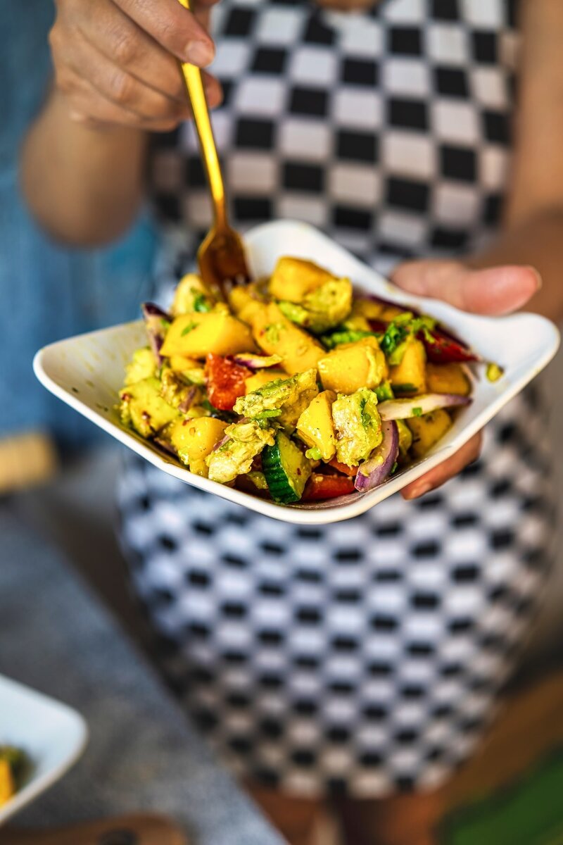 Person serving colorful mango avocado salad with fresh vegetables and herbs in white bowl golden spoon healthy eating clean diet plant based cuisine