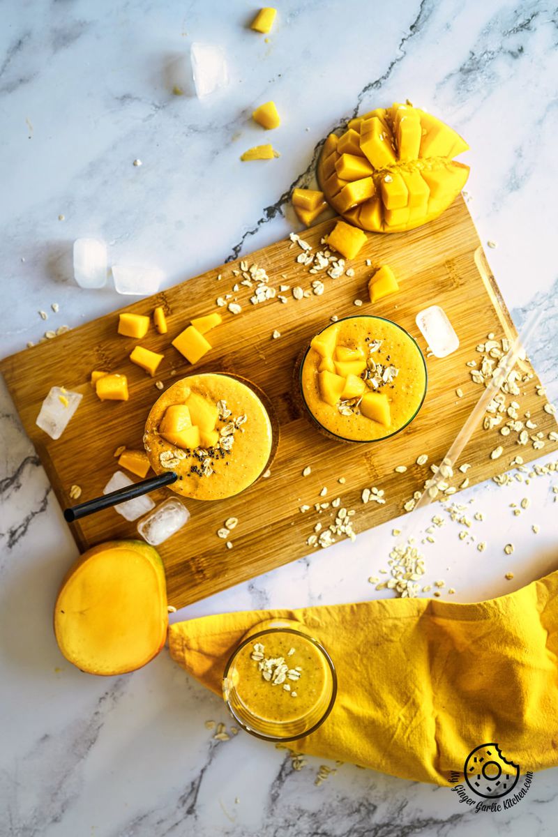 overhead shot of mango oats smoothie in two glasses with a mango cut slice dice, chunks and a yellow napkin