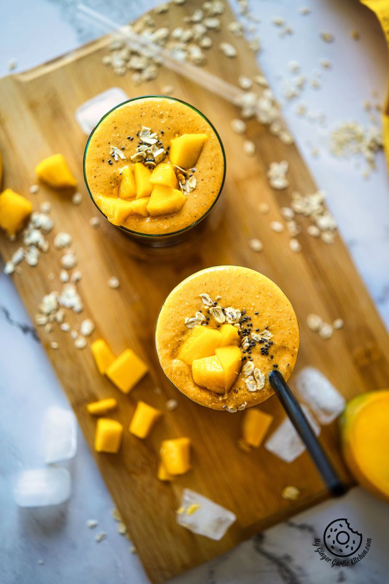 overhead closeup shot of mango oats smoothie in two glasses with ice cubes and mango chunks on a wooden board