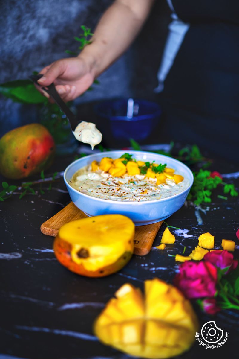 a hand holding a spoon with mango raita over a mango raita bowl