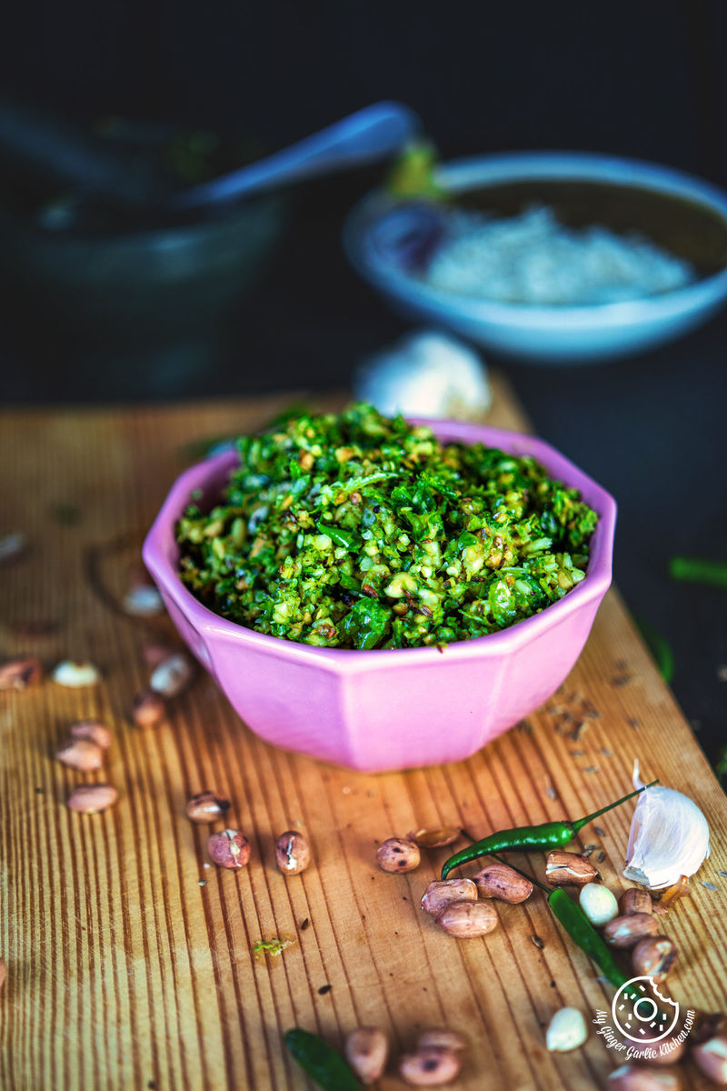 a pink bowl of mirchi ka thecha with garlic cloves, green chilies, and peanuts on side