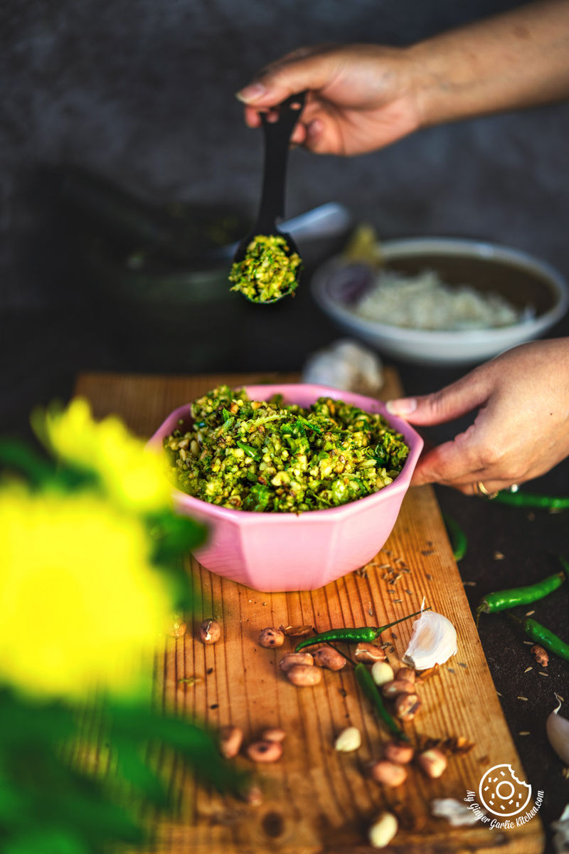 a hand holding a black spoon with mirchi ka thecha over pink bowl with garlic cloves and peanuts on side