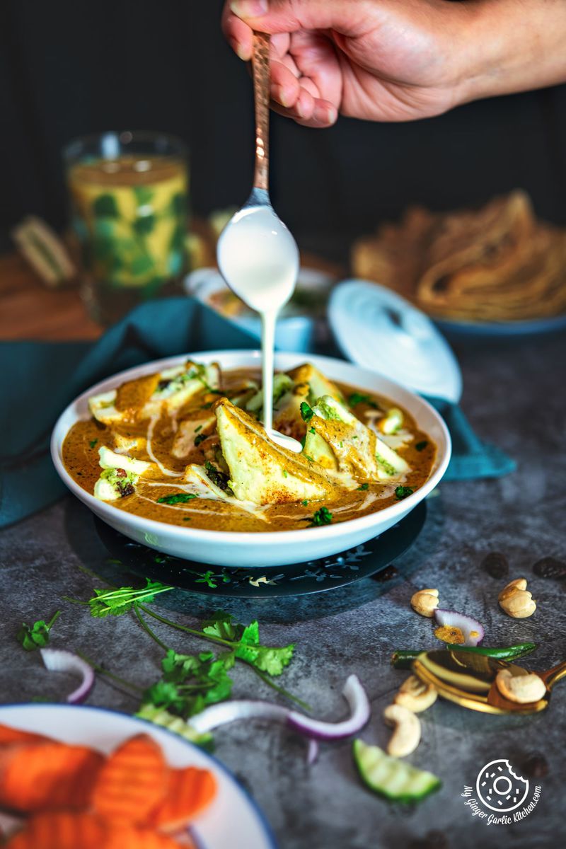a hand drizzling cream oven paneer pasanda served in a white ceramic bowl