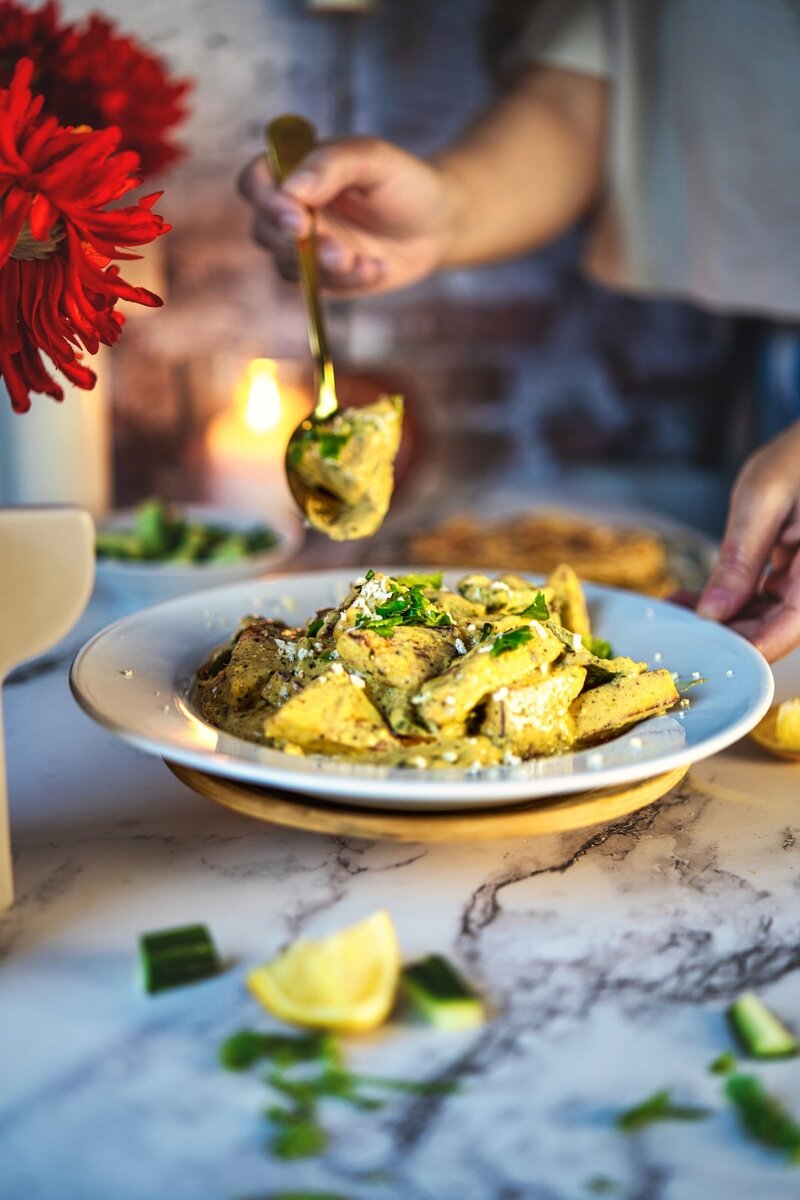 Person serving paneer yakhni from a white bowl, garnished with herbs, with roti, lemon wedges, and salad on a marble table.