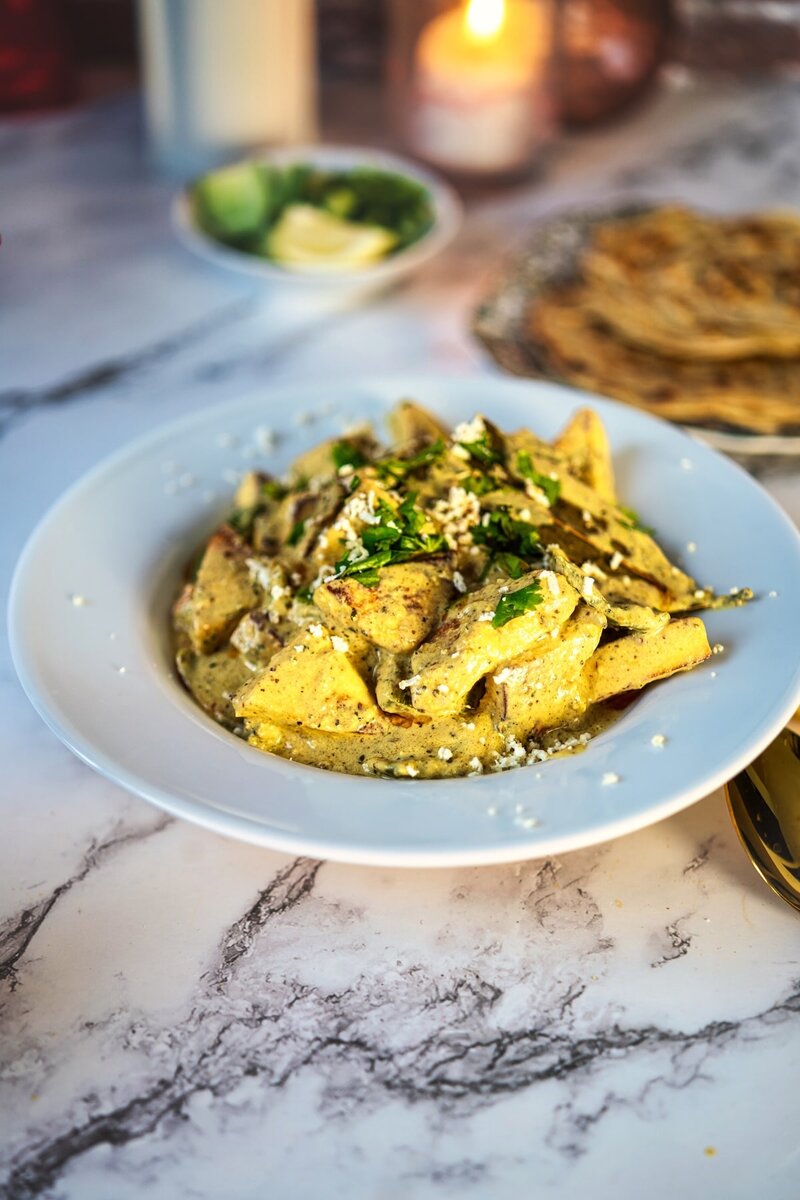 Paneer yakhni served in a white bowl, garnished with fresh herbs and crumbled cheese, with roti and salad in the background on a marble table.