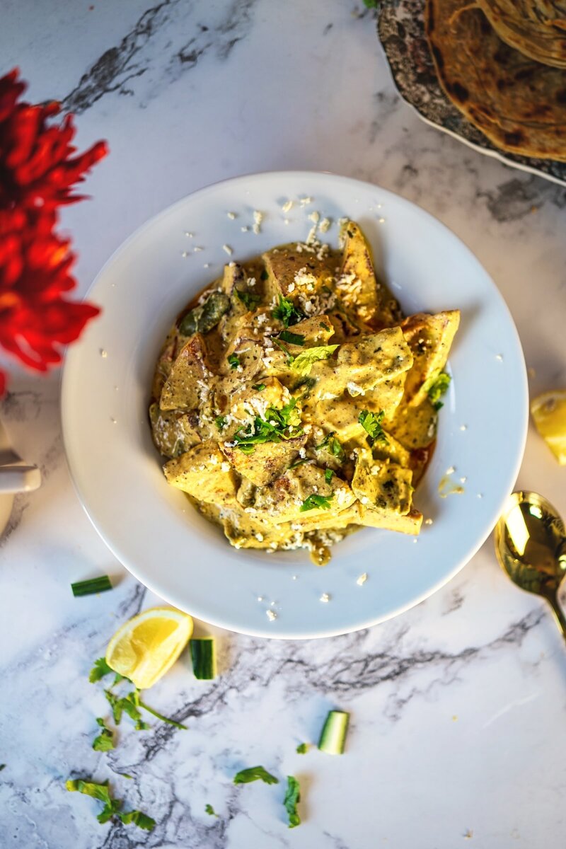 Top view of paneer yakhni in a white bowl, topped with fresh coriander and cheese, surrounded by lemon wedges and cucumber pieces on a marble surface.