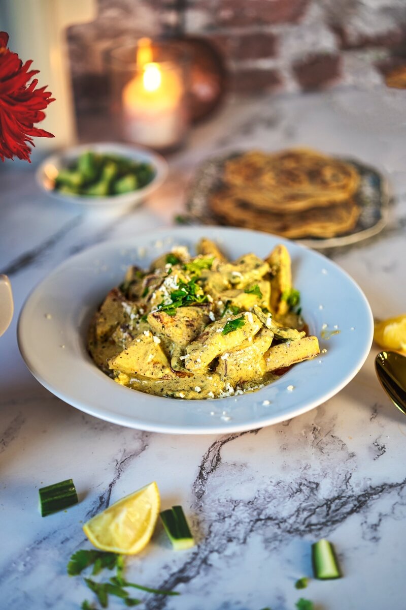Aromatic paneer yakhni curry presented on a blue plate with golden sauce and green herb garnish. Flatbread, cucumber side dish, and a red flower arrangement visible in the background on marble countertop with warm ambient lighting.