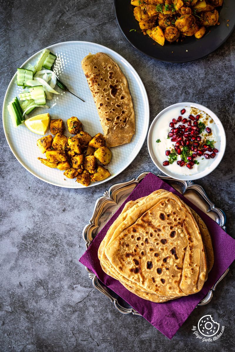 overhead photo of plain triangle paratha on a purple napkin with some raita, sabji and salad