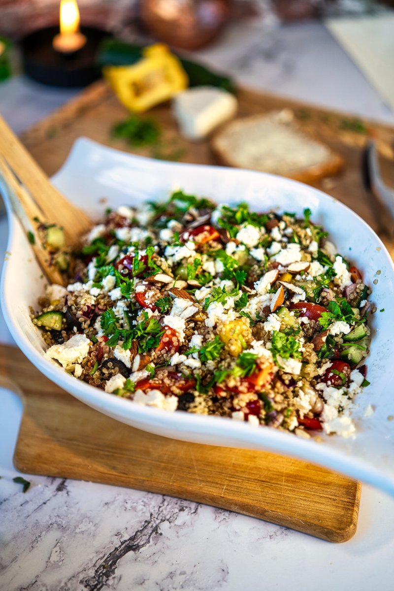 Close-up of a quinoa salad in a white bowl, garnished with cheese, chopped vegetables, and herbs.