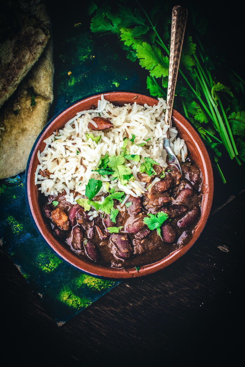 A top-down view of a meal spread including a plate of white rice and kidney bean curry garnished with cilantro and sliced red onions, with two bowls of white rice, a bowl of kidney bean curry, fresh green coriander leaves, and garlic clove on a dark background with a grey cloth.