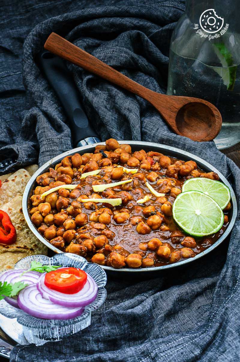 a pan of amritsari chole topped with lemon slices, with a spoon and a glass of water