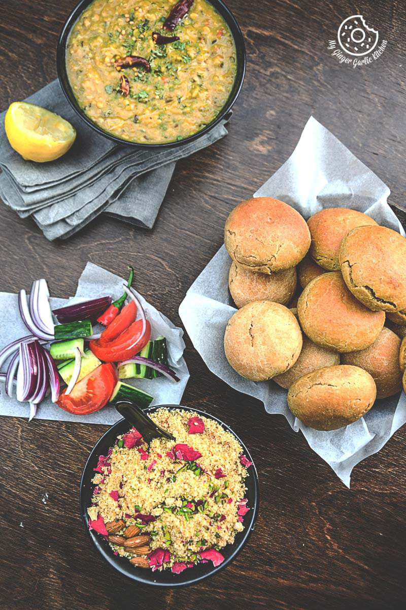 some baati, a bowl of dal, and bowl of churma with fresh salad on a table