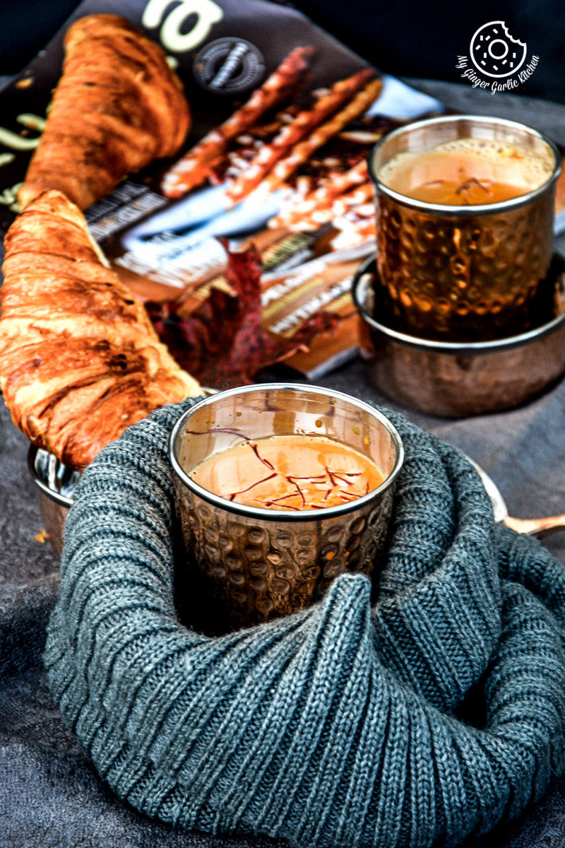 there is a metal cup spiced kesar chai topped with saffron strands and a croissant on a blanket