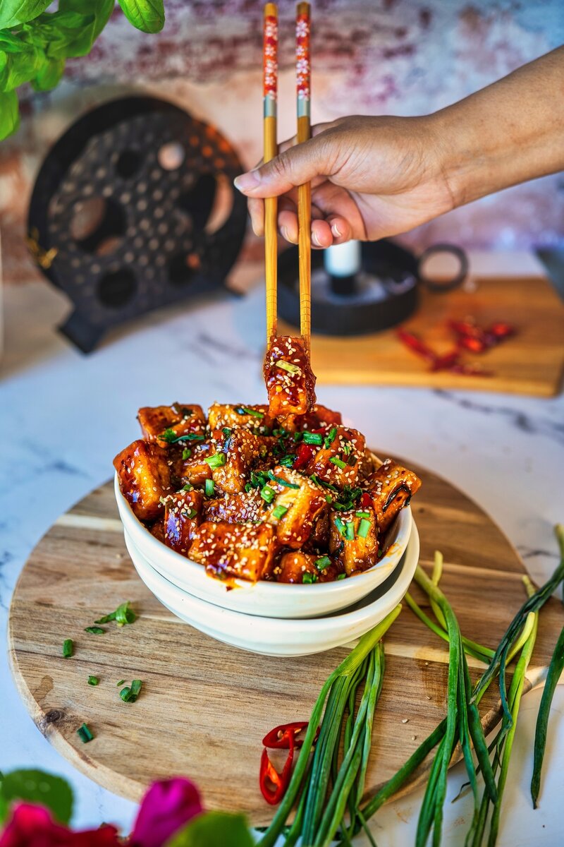 Chopsticks lifting a cube of soy garlic tofu above a bowl, sesame and scallion garnish on rustic kitchen surface