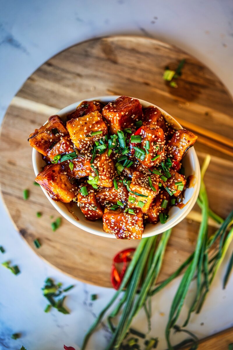 Crispy soy garlic tofu cubes glazed in sticky sauce, topped with sesame seeds and chopped scallions in a white bowl