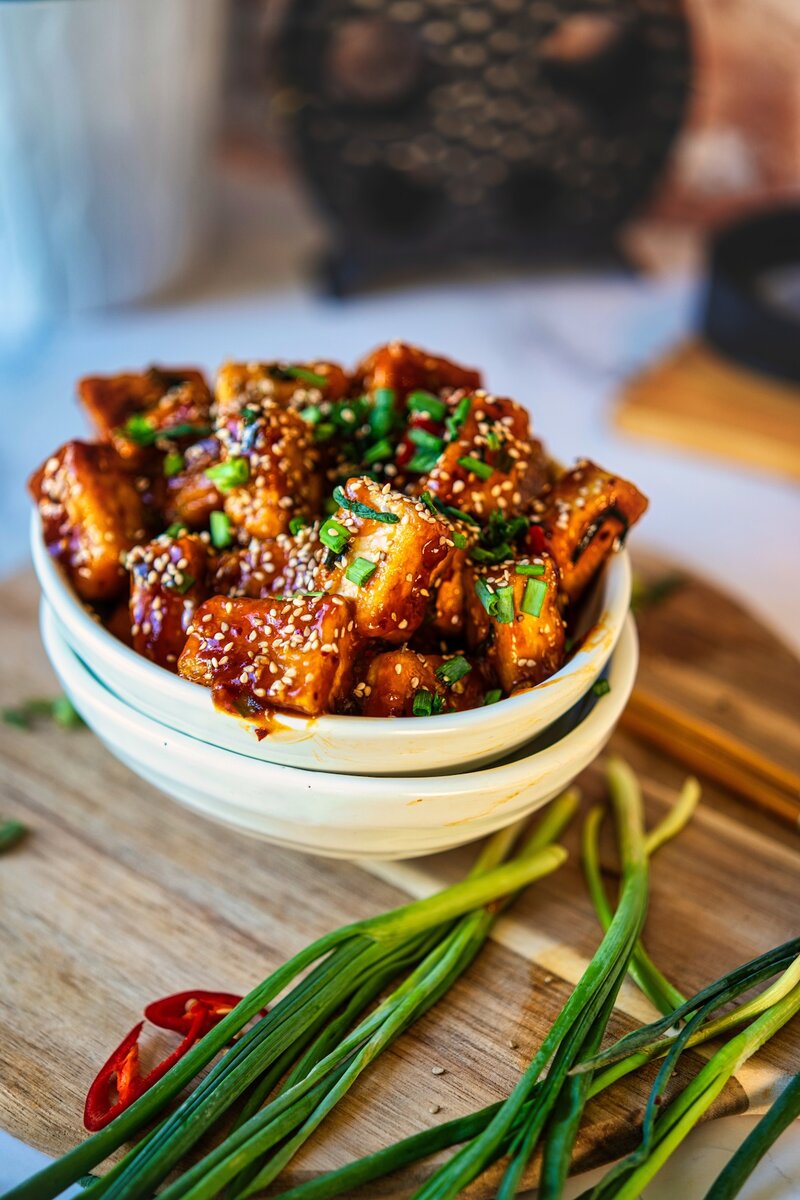 Double-stacked white bowls filled with sticky soy garlic tofu, sesame seeds, and green onions on a wooden board